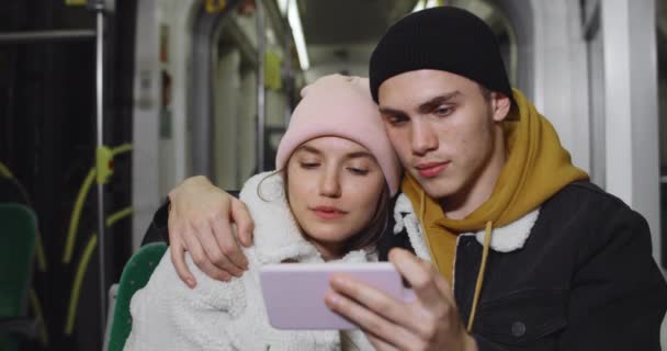 Прекрасная пара смотрит видео на смартфоне, сидя вместе в трамвае. Красивый молодой парень обнимает свою девушку, держа телефон в ландшафтном моде.Концепция реальной жизни. — стоковое видео