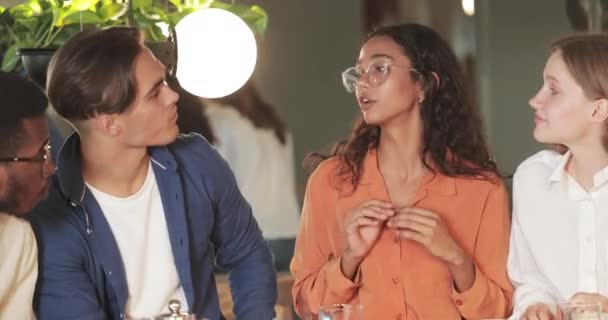 Lächelnde Frau mit Brille im Gespräch mit ihren Freunden in einem modernen Café. Crop Ansicht der multiethnischen jungen Kollegen zuhören Mädchen und lachen. Konzept der Kommunikation und positiver Emotionen. — Stockvideo