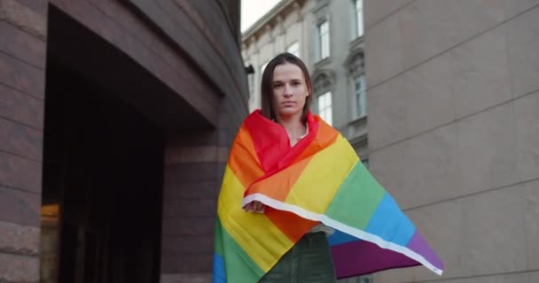 Retrato da jovem mulher cobrindo-se na bandeira do arco-íris enquanto olha para a câmera. Millennial boa aparência menina apoiando movimento lgbt enquanto está em pé na rua da cidade. Ampliar . — Vídeo de Stock