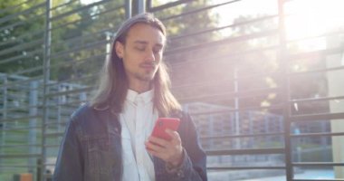 Uzun saçlı genç adam spor sahasının yanında güneş ışığında beklerken akıllı telefondan sohbet ediyor. Küpeli ve sakallı telefon kullanan bin yıllık bir adam. Gerçek hayat kavramı.