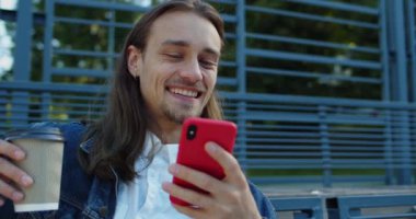 Telefon ekranına bakarken elinde kağıt kahve ve kahkaha bardağı tutan mutlu sakallı adamın yakın görüntüsü. Dışarıda bankta otururken cep telefonu kullanan uzun saçlı genç adam..