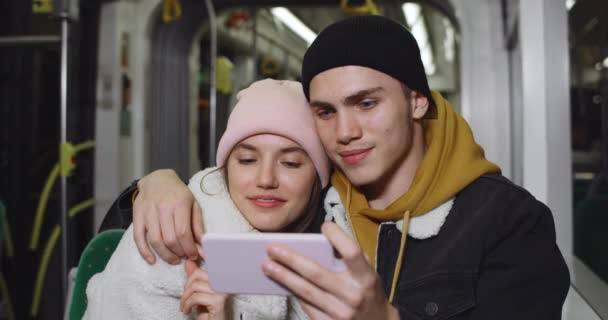 Millennial pár sledování legrační video na smartphonu a směje se, zatímco sedí spolu v autobuse. Mladý muž objímající jeho hezká přítelkyně při držení telefonu v režimu krajiny. — Stock video