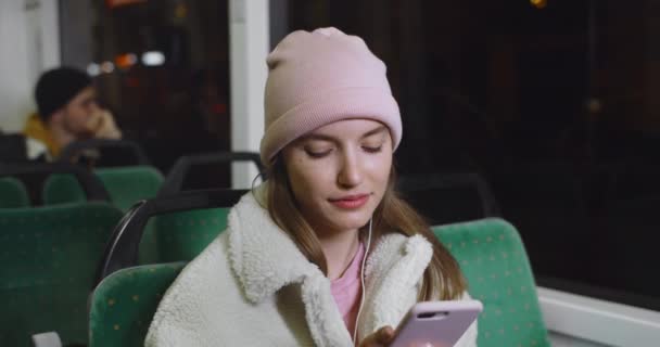 Bin yıllık güzel bir kadının toplu taşıma aracına binerken akıllı telefonunu kullanması. Kulaklıklı güzel kız internette gezinirken gülümsüyor ve telefon ekranına bakıyor.. — Stok video