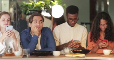 Gençler kafede birlikte vakit geçirirken telefonlarını kullanıyorlar. Yakışıklı sıkılmış adam, akıllı telefonları olan ve yemek yiyen çok ırklı arkadaşlar arasında oturuyor..