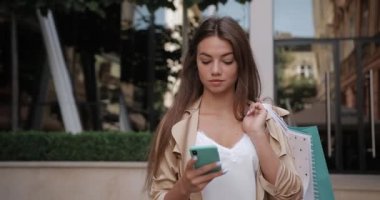 Neşeli esmer kadının haberlerde kayarken telefon ekranına bakması ön planda. Millennial çekici kız, kağıt torbalar taşırken ve sokakta yürürken gülümsüyor..