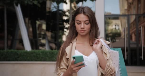 Vista frontale della donna bruna gioiosa che guarda lo schermo del telefono mentre scorre il feed di notizie. Millennial ragazza attraente sorridente mentre trasporta sacchetti di carta e camminando per strada della città. — Video Stock