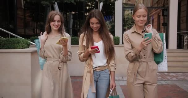 Trzy radosne młode kobiety korzystające ze smartfonów podczas spaceru po ulicy miejskiej. Atrakcyjne dziewczyny z papierowymi torbami uśmiechnięte patrząc na ekrany telefonu po zakupach. — Wideo stockowe