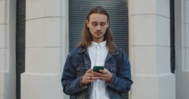 Küpeli hippi genç adam akıllı telefonunu kullanırken yana bakıyor. Sakallı milenyum sakallı adam günlük giysiler giyiyor. Şehir caddesinde dikilirken yazıp internette geziniyor..