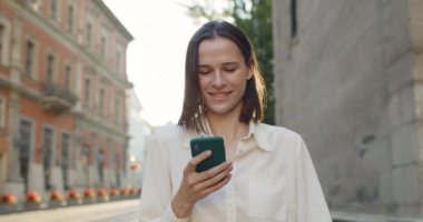 Dışarıda yürürken cep telefonu kullanan mutlu genç bir kadının yakın görüntüsü. Şehir caddesinde kaydırırken ve telefon ekranına bakarken gülümseyen güzel kız. Gerçek hayat kavramı.