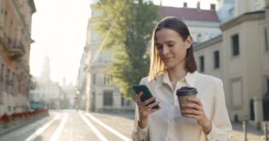 Çekici genç bir kadın telefonda güzel haberler veriyor ve elinde kahve fincanı tutarken gülümsüyor. Şehrin caddesinde dikilirken akıllı telefonunu kullanan neşeli kızın yan görüntüsü.