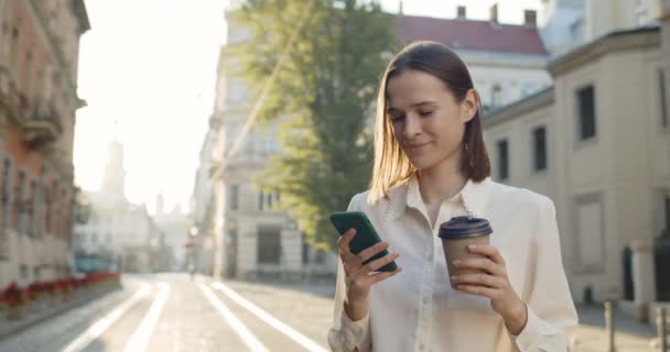 Attraktiv ung kvinna som har goda nyheter på telefon och ler medan du håller kaffekopp utomhus. Sidovy av glad flicka som använder sin smartphone när du står på stadens gata. — Stockvideo