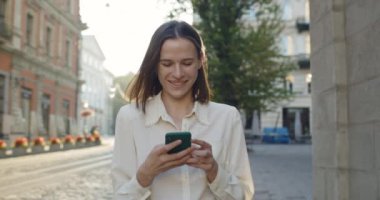 Akıllı telefon ekranına bakıp eski şehir caddesinde yürürken gülümseyen mutlu genç kız. Çekici bir milenyum kadınının açık havada telefon kullanırken keyifli görünümü..
