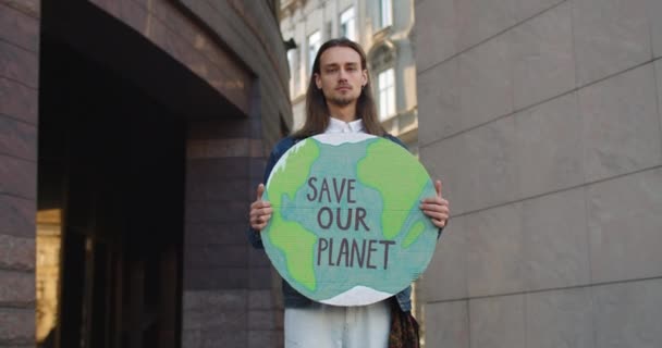 Portret van jonge mannelijke eco-activist met oorbellen die naar de camera kijken terwijl ze op straat staan. Serieuze millennial man die het Aardemodel vasthoudt met redding van onze planeet schrijven. Inzoomen.. — Stockvideo