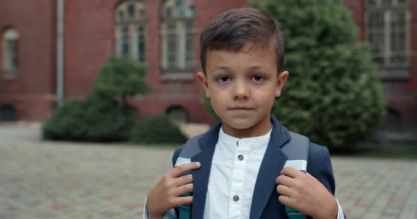 Zbliż widok dzieciaka z torbą patrzącego w kamerę. Słodki chłopak w szkolnym mundurze, pozujący na zewnątrz. Pojęcie edukacji i dzieci. — Wideo stockowe