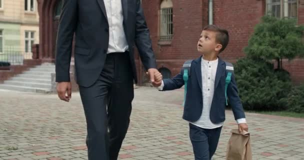 スーツを手に手をつないで袋と彼の父と小さな男の子の作物ビュー。小さな子供と父は学校の庭を歩いて通信します。教育と子供の概念. — ストック動画