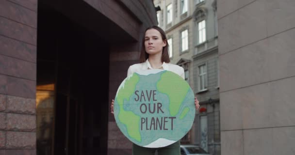 Kobieta eko-aktywistka odwracając głowę i patrząc w kamerę stojąc na ulicy.Młoda ładna kobieta trzyma kartonowy plakat z zapisać naszą planetę pisania. — Wideo stockowe