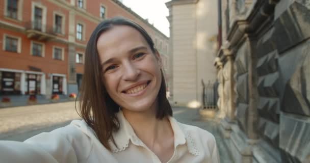 Joven mujer atractiva teniendo video conversación y sonriendo. Mujer feliz usando el teléfono inteligente para la comunicación mientras está de pie en la calle de la ciudad vieja. — Vídeos de Stock