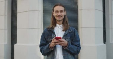Küpeli, başını kaldırıp kameraya bakan neşeli genç adamın portresi. Bin yıllık uzun saçlı adam akıllı telefon kullanıyor ve caddede dururken gülümsüyor. Yakınlaştır.
