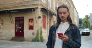 Millennial hipster 'lı adam sosyal medyada haber yayını yaparken akıllı telefonunu kullanıyor. Uzun saçlı ve küpeli sakallı adam. Şehir caddesinde yürürken mesaj yazıyor..