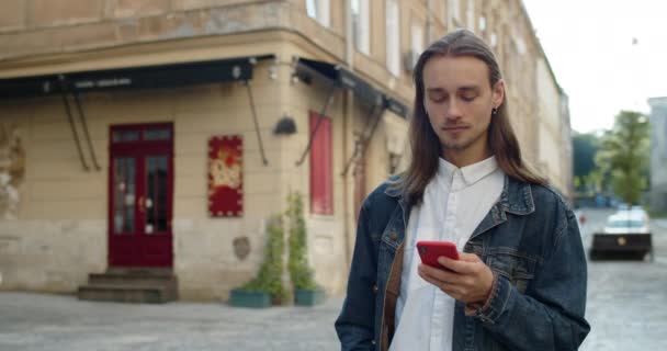 Millennial hipster 'lı adam sosyal medyada haber yayını yaparken akıllı telefonunu kullanıyor. Uzun saçlı ve küpeli sakallı adam. Şehir caddesinde yürürken mesaj yazıyor.. — Stok video