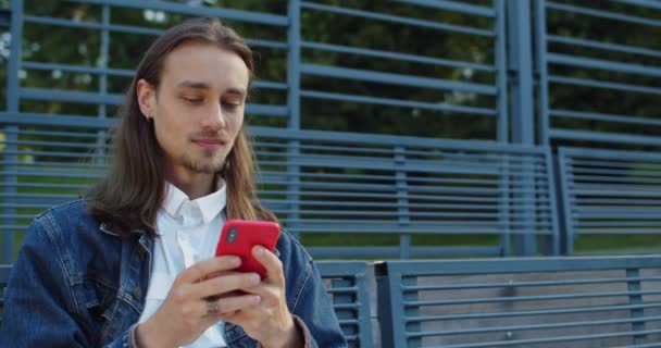 Close up visning af glade unge mand med øreringe har gode nyheder, mens du bruger smartphone. Skægget tusindårig fyr ser på telefonens skærm og smiler mens du sidder på bænken udendørs. – Stock-video