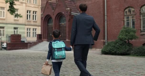 Rückansicht von Papa im Anzug und seinem kleinen Sohn mit Tasche, der am ersten Schultag zur Schule geht. Der Vater hält die Hand eines kleinen Kindes, während er auf der Straße geht. Konzept von Bildung und Kindern. — Stockvideo