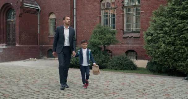 Snygg man i kostym och litlle son med ryggsäck kommunicerar samtidigt hålla hand i hand. Far med ett barn som går på skolgården. Begreppet utbildning och barn. — Stockvideo