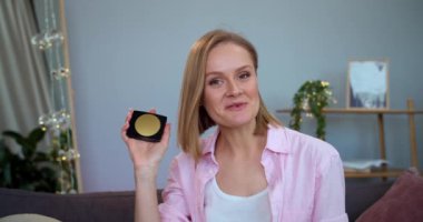 Neşeli genç bir kadın, evdeki koltukta otururken kameranın kozmetik paletine konuşuyor ve onu gösteriyor. Ürün değerlendirmesi yapıyor. Güzellik ve vloglama kavramı.