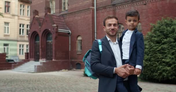 Porträtt av glad ung far och hans bedårande son tittar på kameran och ler. Snygg man i kostym som håller pojken och hans väska stående på skolgården. Begreppet familj. — Stockvideo