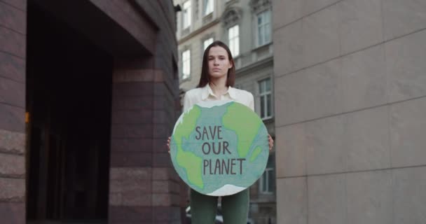 Porträt einer jungen Öko-Aktivistin, die auf der Straße den Kopf dreht und in die Kamera blickt. Ernstes Millennial-Mädchen mit einem Pappschild mit der Aufschrift "Rettet unseren Planeten". Vergrößern. — Stockvideo
