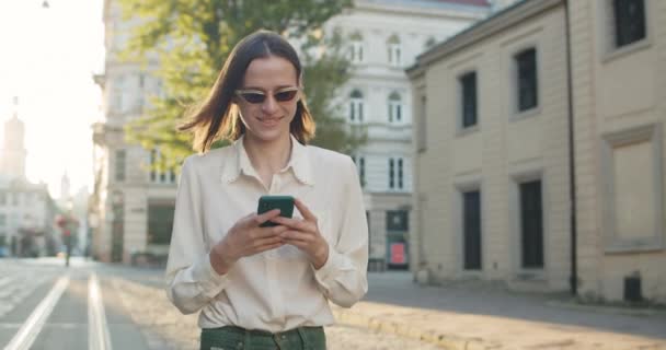 Glada kvinna i elegant dagsljus tittar på telefonskärmen när du går på stadens gata. Millennial kvinna skriva meddelande och ler när du använder sin smartphone. Begreppet verklighet. — Stockvideo