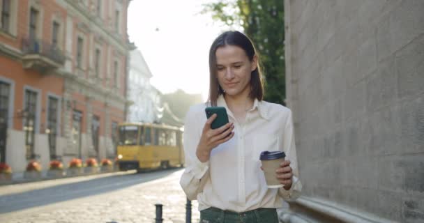 Schöne Frau mit Smartphone und Kaffeetasse, während sie an der Altstadtstraße steht. Fröhliches Mädchen, das auf den Handybildschirm schaut und beim Surfen im Internet lächelt. Straßenbahn im Hintergrund. — Stockvideo