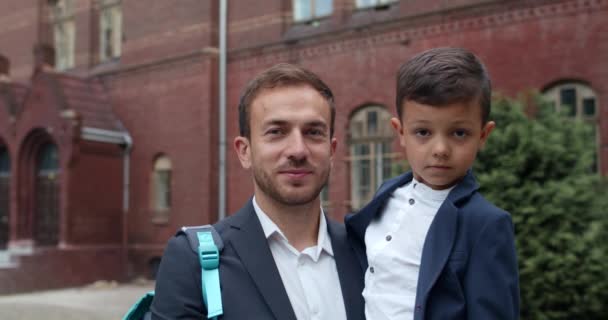 Närbild av glad pappa och hans söta son tittar på kameran och ler. Ung man i kostym håller liten skolpojke och hans ryggsäck när han står på gatan. Begreppet familj. — Stockvideo