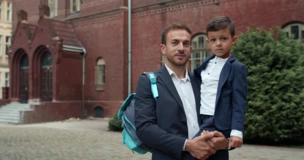 Porträtt av lycklig ung far och hans söta son vrider huvudet och tittar på kameran. Le kille i kostym hålla liten pojke och hans ryggsäck när du står på skolgården. — Stockvideo