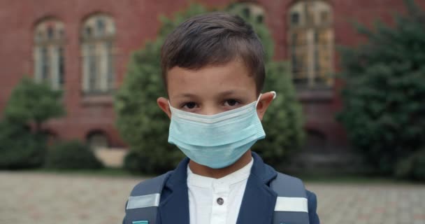 Porträtt av pojke klädd i medicinsk skyddsmask tittar mot kameran. Barn med ryggsäck bär uniform poserar när du står nära skolan. Begreppet barn, utbildning och virus. — Stockvideo