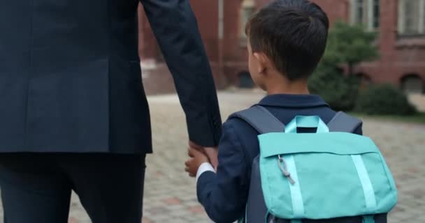 Närbild på pojken med väskan som tar faderns hand och de går till skolan. Man i kostym väntar på sin son när han står på gården. Begreppet utbildning och familj. — Stockvideo