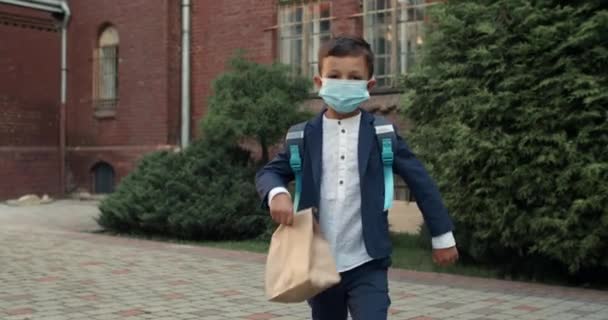 Beskär syn på liten unge med skyddande medicinsk mask på ansiktet promenader och tittar till kameran. Pojke i uniform med ryggsäck och lunch på väg till skolan. Begreppet virus. — Stockvideo