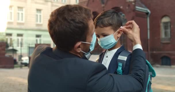 Närbild av kärleksfull far bär medicinsk mask på sin son ansikte när du skickar honom till skolan. Pojke med ryggsäck ger hög fem och kör medan man i kostym står och tittar på. — Stockvideo