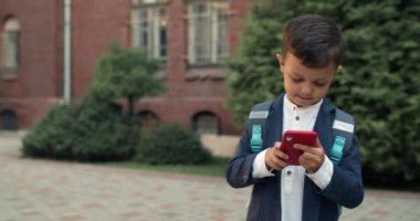Sokakta yürürken telefon ekranına bakan sevimli genç bir çocuk görüntüsü. Sırt çantalı çocuk akıllı telefon kullandığı için okul üniforması giyiyor. Teknoloji ve çocuk kavramı.