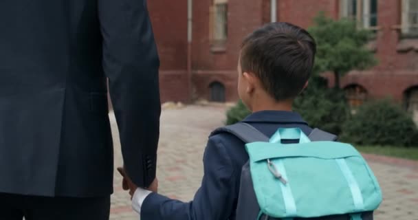 Vista trasera de un niño pequeño con la mochila cogida de la mano de su padre en traje. Hombre abrazando a su hijo mientras lo lleva a la escuela. Concepto de educación y familia. — Vídeo de stock