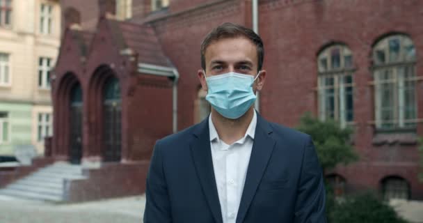 Tıbbi koruyucu maske takan ve kameraya bakan genç bir adamın portresi. Takım elbiseli bir erkek işadamı sokakta dikilirken poz veriyor. Halk, eğitim ve virüs kavramı. — Stok video