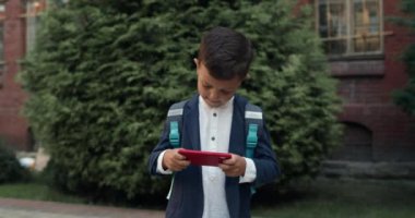 Küçük çocuk sokakta yürürken akıllı telefondan oyun oynuyor. Okul yolunda üniformalı bir çocuk telefon ekranında yemek yiyor. Oyun ve çocuk kavramı.