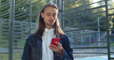 Spor sahasının yanında dururken akıllı telefon kullanan sakallı bir gencin görüntüsü. Küpeli ve sakallı hippi adam internette geziniyor ve ileriye bakıyor. Gerçek hayat kavramı.