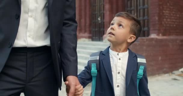 Närbild av liten pojke med ryggsäck tittar och prata med sin far när du går på gatan. Addorable kid ger high five till pappa samtidigt som han håller handen på väg till skolan. — Stockvideo