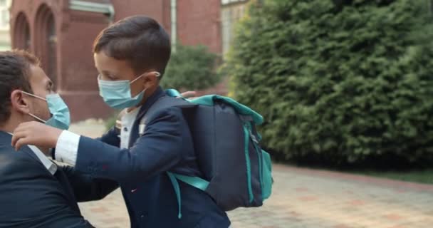 Αγαπώντας τον μπαμπά και το μικρό παιδί φορώντας ιατρικές μάσκες αγκαλιασμένοι στην αυλή του σχολείου. Νεαρό αγόρι με τσάντα να κάνει κόλλα πέντε και να τρέχει ενώ ο κουστουμάτος στέκεται και προσέχει τα νώτα του. Έννοια του ιού. — Αρχείο Βίντεο
