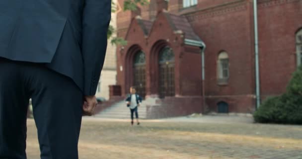 Muž v obleku čeká na svého syna po vyučování poblíž školní budovy. Usmívající se chlapeček s taškou v uniformě, jak utíká do otcova objetí. Pojem rodina a vzdělávání. — Stock video