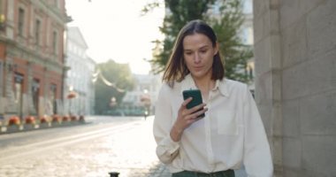 Genç, neşeli bir kız sokakta telefon ekranına bakarken şaşırmış yüz ifadesi takınıyor. Güzel milenyum kadını, akıllı telefonundan iyi haberler alırken seviniyor ve gülümsüyor..