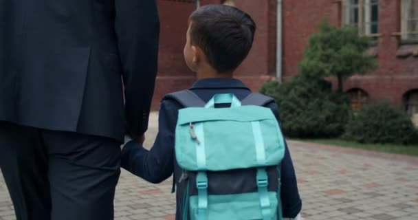 Πλάτη άποψη του μικρού παιδιού με τσάντα κρατώντας το χέρι στο χέρι ο πατέρας του στο κοστούμι. Άντρας πηγαίνει το γιο του στο σχολείο. Συγκεντρώσου στο αγόρι. Έννοια της εκπαίδευσης και της οικογένειας. — Αρχείο Βίντεο
