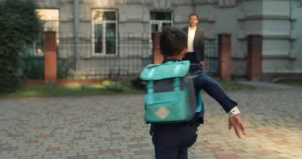 Baksidan av en liten pojke med ryggsäck springande i sin far omfamna. Glada unga man väntar sin son på skolgården efter första dagen av studierna. Begreppet lycklig familj. — Stockvideo
