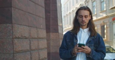 Eski şehir caddesinde yürürken akıllı telefon ekranına bakan mutlu genç adam. Küpeli sakallı ve gülümseyen binlerce yıllık adamın telefon kullanırken görüntüsü..